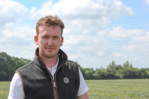 A photo of Liam Wilkinson, Head of Technical for Limagrain Field Seeds UK. Liam is stood to the left hand side and he is in a field with crops grown in the background. He is wearing a Limagrain Field Seeds UK gilet, with a bright blue sky and white clouds in the background.